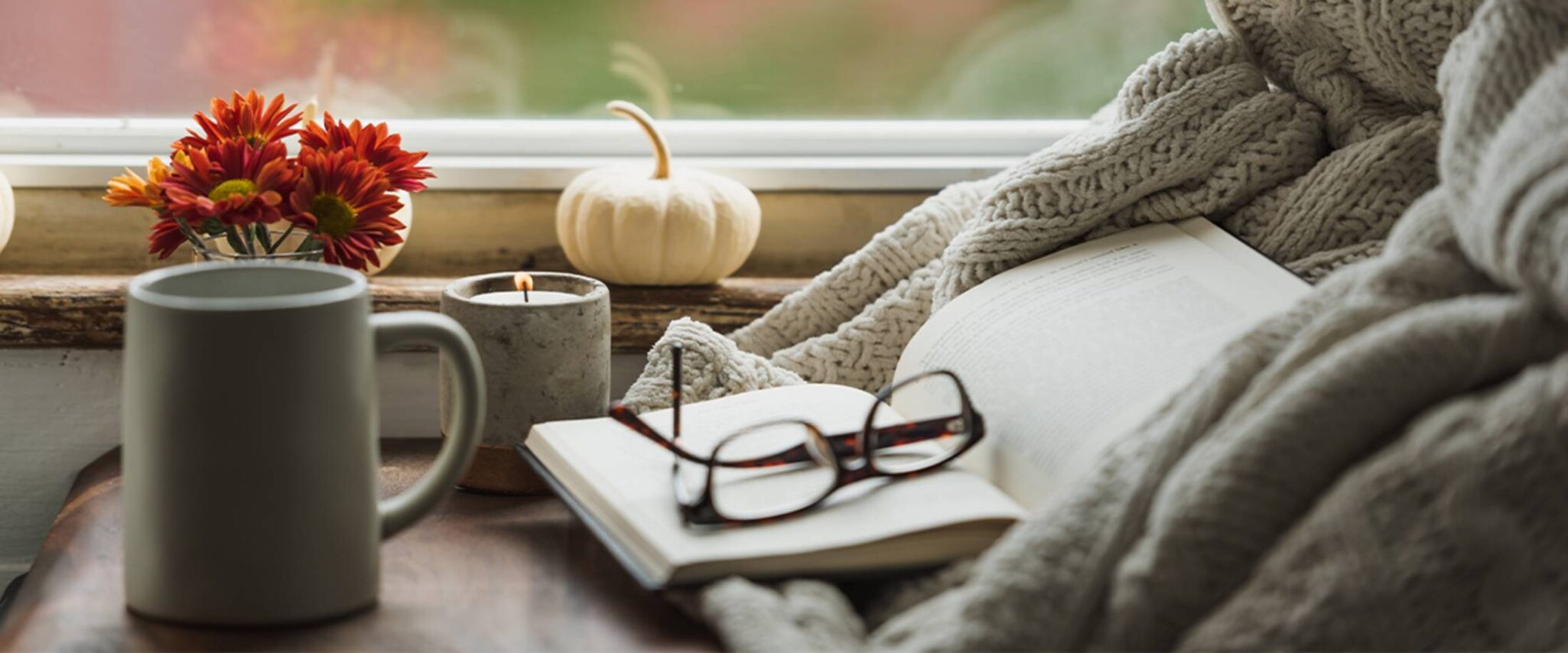 close up of reading glasses laying on an open book surrounded by a candle, coffee mug, flowers, and a small white pumpkin