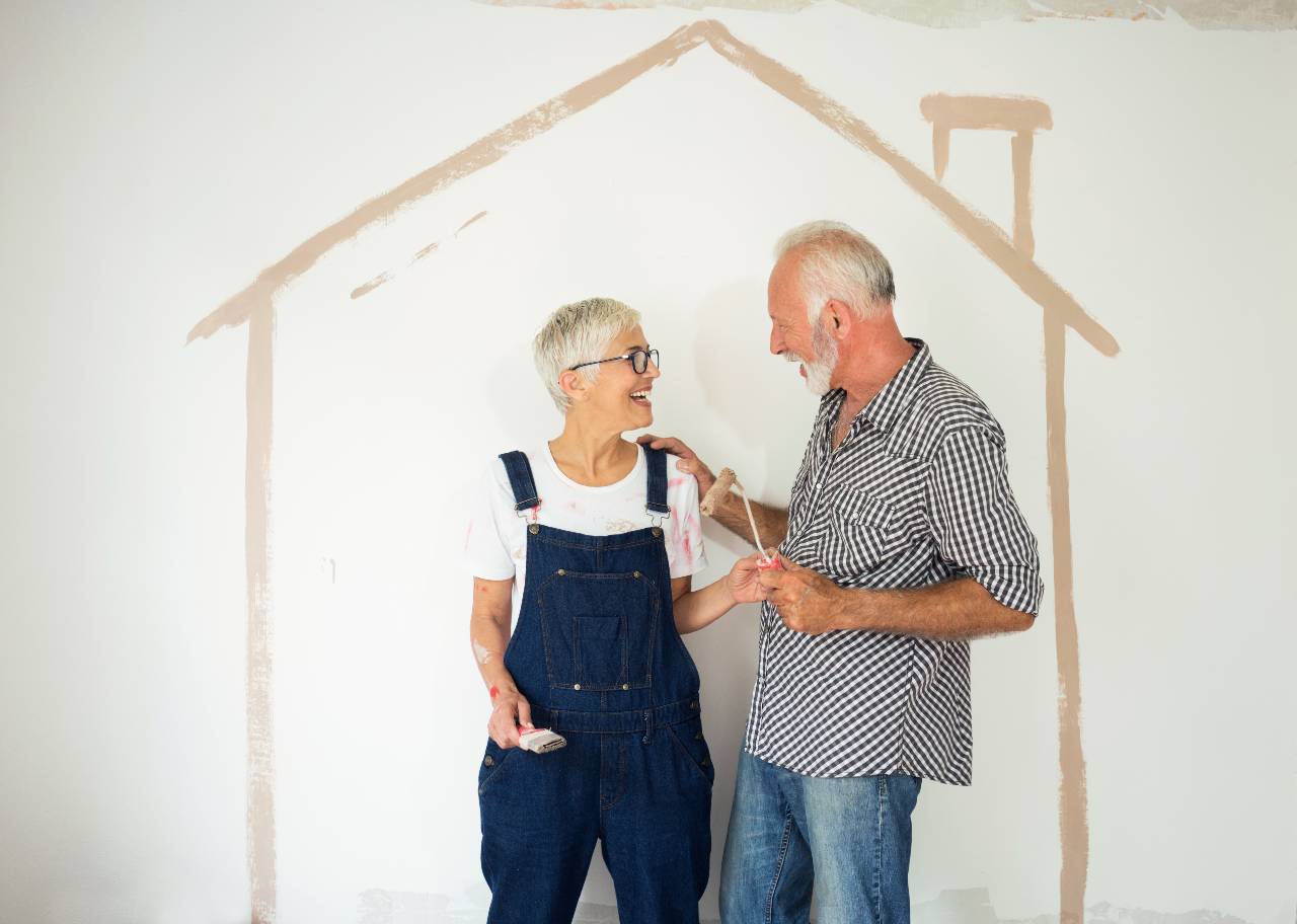 A man and a woman in painting clothes stand in front of a while wall with a painted outline of a house on it