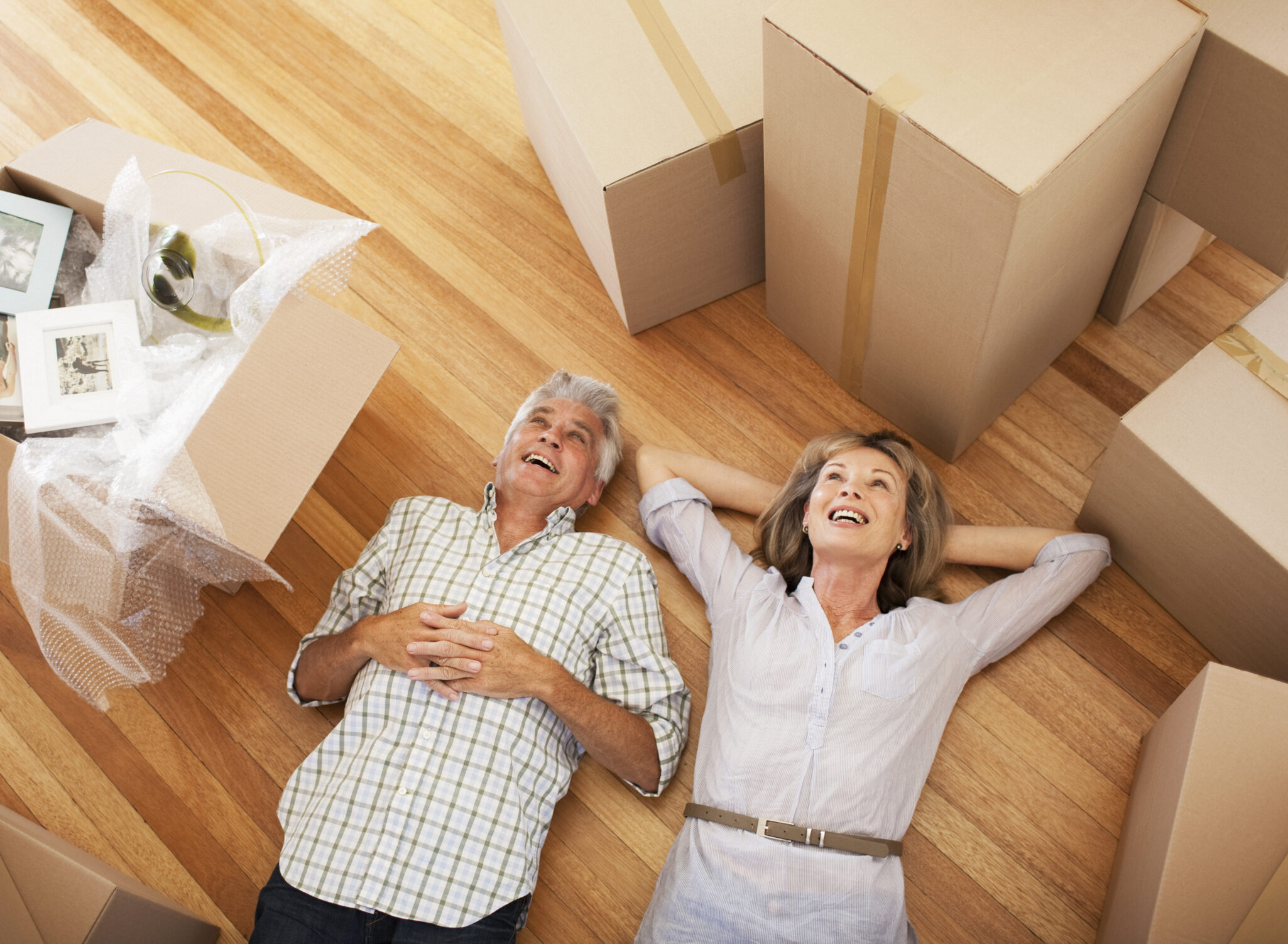 A couple lay on the ground and look up at the ceiling, surrounded by moving boxes