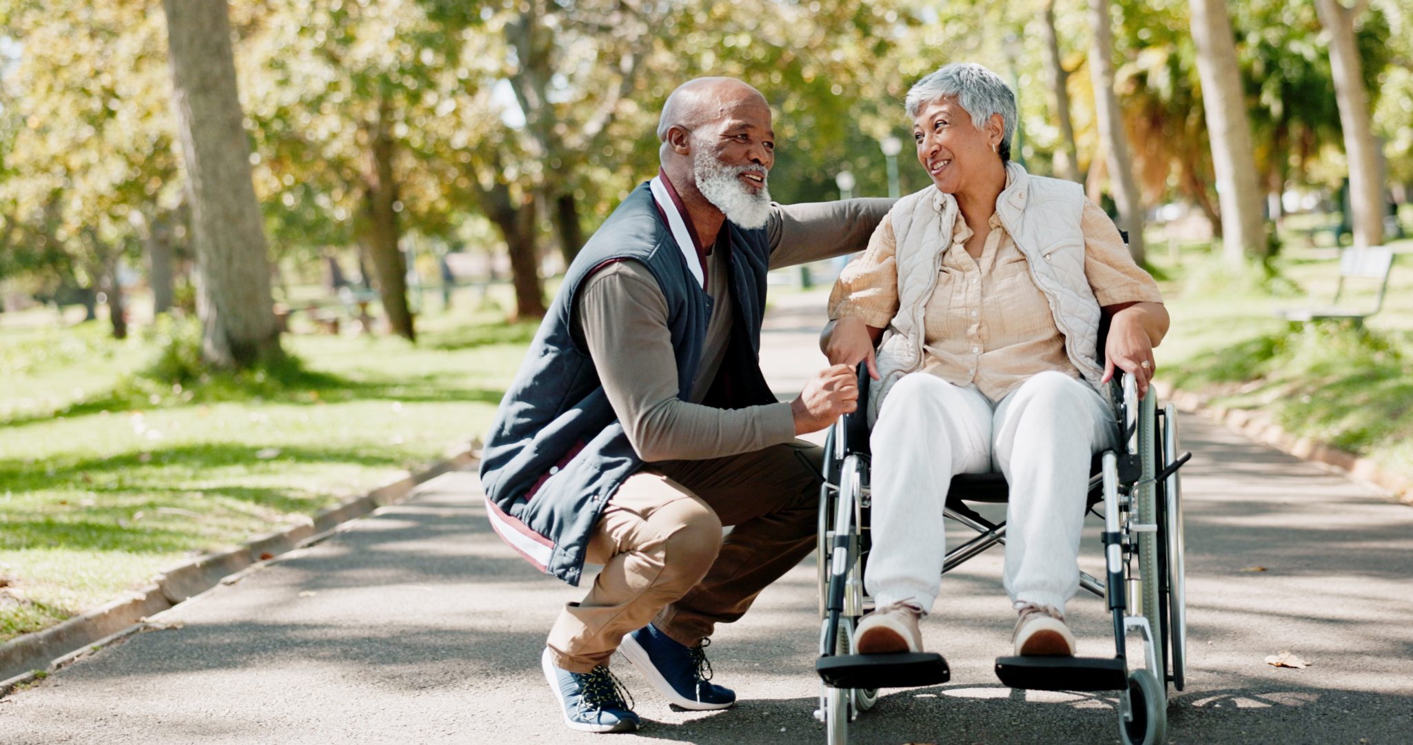 Senior man smiling at senior woman in wheelchair while on walk in park