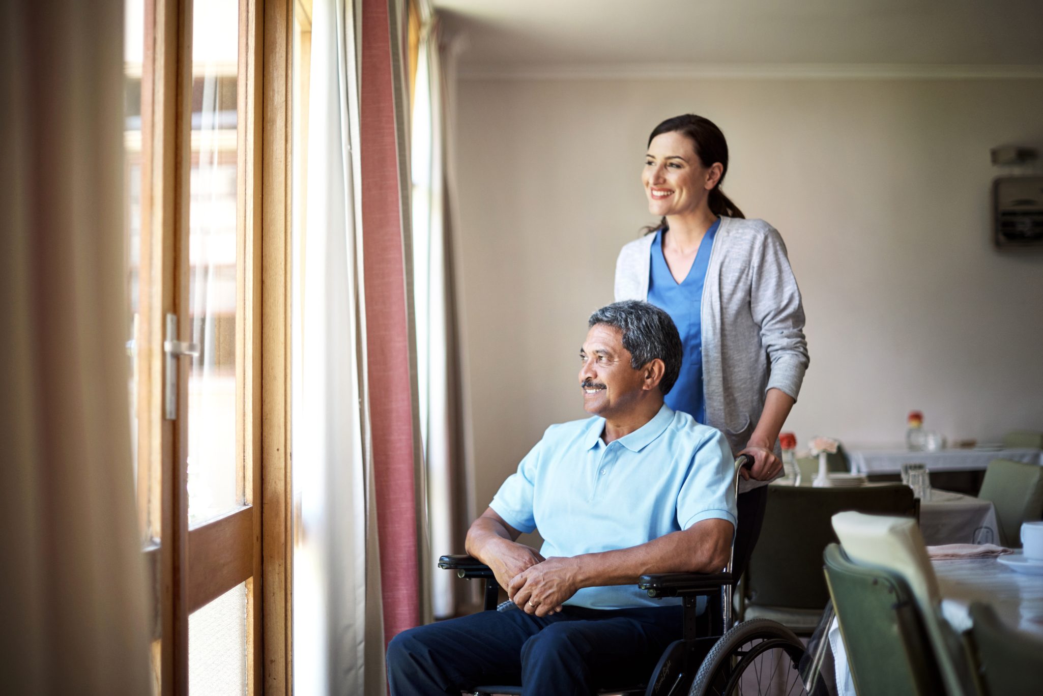 Senior man in wheelchair looking out window with female nurse behind him